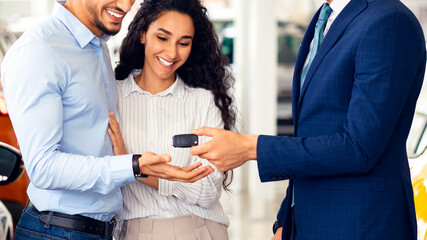 Smiling sales manager giving happy young middle eastern family key from brand new auto, side view. Emotional arabic couple buying car at luxury dealership salon. Car purchasing concept