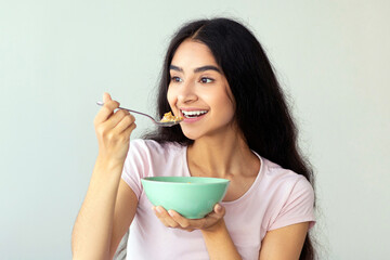 Attractive Indian lady eating yummy cereal with milk while sitting on comfortable bed at home, full...