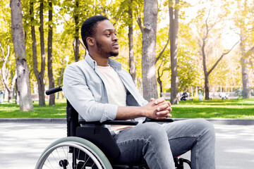 Fototapeta premium Full length of peaceful black handicapped guy in wheelchair having walk at urban park in autumn. Paraplegic African American man deep in thought, spending time outdoors in fall