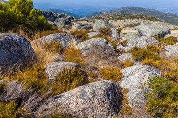 A rugged mountain landscape with large boulders, sparse vegetation, and a distant view under a clear sky