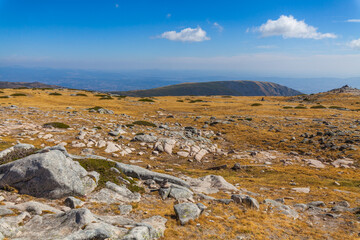 A high-altitude mountain landscape with rocky outcrops, dry grass, and a distant panoramic view under a blue sky with scattered cloudsё