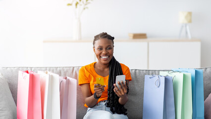 Shopping from home. Joyful black woman with credit card and smartphone confirming her online order,...