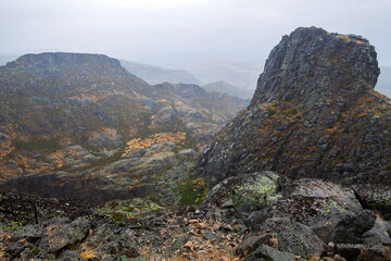 A rugged mountain landscape with rocky outcrops and sparse vegetation is shrouded in mist and low clouds