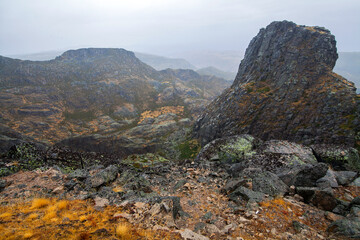 A rugged mountain landscape with rocky outcrops and sparse vegetation is shrouded in mist and low clouds