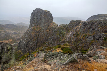 A rugged mountain landscape with rocky outcrops and sparse vegetation is shrouded in mist and low clouds