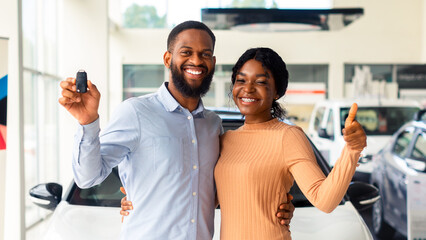 Happy Black Couple Holding Keys From New Car And Showing Thumb Up, Cheerful Young African American Spouses Purchased Automobile In Dealer Showroom, Looking And Smiling At Camera, Free Space