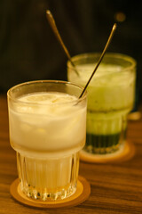 A cozy close-up of iced lemonade and matcha drinks served in elegant glasses with spoons, placed on coasters atop a wooden table. Warm lighting adds a comforting ambience.