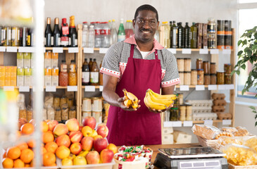 While working in store, African man stand at sales area, ready to communicate with customers. He in apron waiting for orders, standing near glass display case with alcohol and sweet