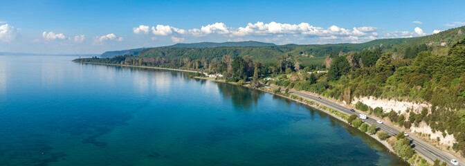 Fototapeta premium Highway winds along a scenic Lake Taupo shore, surrounded by lush greenery. Tranquil waters reflect the sky. Beautiful New Zealand landscape. LAKE TAUPO, TURANGI, WAIKATO, NEW ZEALAND