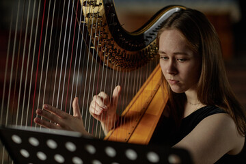 Portrait of young thoughtful woman looking at notes playing harp softly while performing classical music on stage in dim light