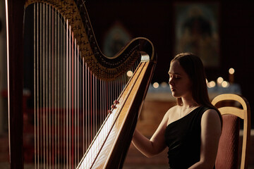 Side view of young female musician in elegant dress sitting at harp, preparing to perform classical music in church with contrasting light casting on instruments strings