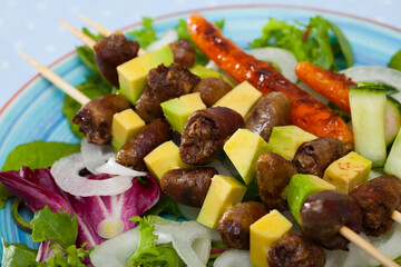 Closeup of grilled chicken hearts on skewers with avocado, baked carrots and fresh greens on colored plate