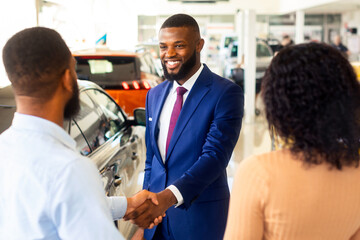 Obraz premium Car Dealership Concept. Salesperson Handshaking With Young Black Customers Couple In Showroom, African American Spouses Buying New Automobile In Modern Auto Salon, Selective Focus On Manager