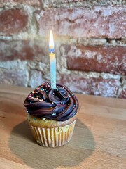 A single vanilla cupcake topped with rich, swirled chocolate frosting. Multicolored sprinkles add a festive touch, while a single lit candle stands proudly at the center.