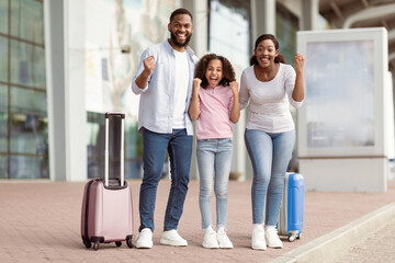 Fototapeta premium Holiday Trip After Lockdown. Full body length portrait of excited African American mum, daddy and daughter going on a journey. Family shaking clenched fists, making win gesture, standing near airport