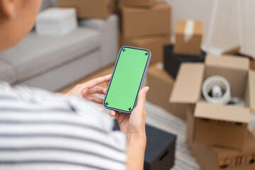 Tech-Savvy Move: Woman holds a smartphone with a green screen amid blurred cardboard boxes in a...