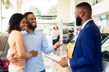 Obraz premium Family Buying Car. African American Spouses Purchasing New Vehicle In Dealership Center, Happy Black Couple Discussing Contract Details With Smiling Salesman In Modern Showroom, Closeup Shot
