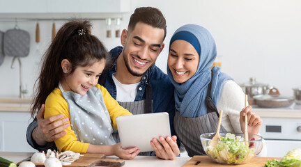 Happy Muslim Family Using Digital Tablet While Cooking In Kitchen Together, Cute Little Arab Girl And Her Islamic Parents Checking Online Recipe On Tab Computer While Preparing Food At Home