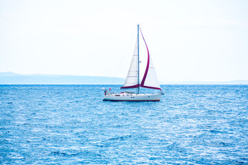 Sailboat gliding across the calm ocean waters under a clear blue sky. Yacht with white sails vast expanse of blue water. Background with faint outline of distant land, creating a serene maritime scene