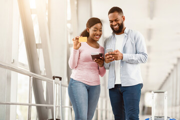Happy Loving Couple In Airport. Portrait of smiling black man and woman using mobile phone and holding golden debit credit card with passports and tickets, standing at terminal with luggage