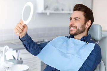 Male patient looking at his beautiful healthy white smile, holding dental hand mirror, happy young...