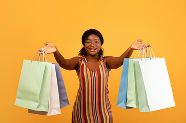 Seasonal sales and shopping concept. Charming young black woman showing bright shopper bags with purchases at camera on orange studio background. Consumerism, special offers and discounts