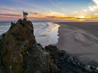 Coastal sunset view of a rocky outcrop with a solar-powered observation tower. Gentle waves lap the shore. Tranquil scene. WHATIPU, AUCKLAND, NEW ZEALAND