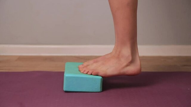 Slim female bare feet are doing foot exercises lifting raise on toes up and down on heels on yoga block on mat indoors at home interior. Foot, body care. Training to correct feet using brick. Closeup