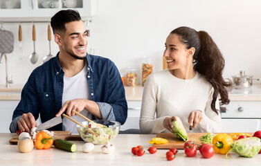 Young Arab Spouses Cooking Healthy Lunch Together In Kitchen, Cheerful Romantic Middle Eastern Lovers Making Fresh Vegetable Salad, Looking And Smiling To Each Other While Preparing Food, Free Space