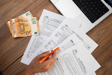Obraz premium Man filling out tax forms with euro banknotes and a laptop on a wooden desk. Financial planning and tax preparation concept. Ideal for accountants and tax season visuals 