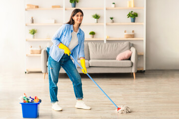 Chores Concept. Portrait of casual woman washing hardwood laminate flooring using string mop pad, standing near bucket with cleaning agents and detergents, posing and looking at camera