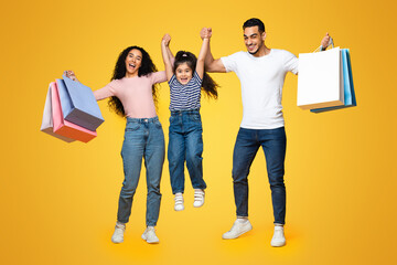 Fototapeta premium Big Sales. Overjoyed Middle Eastern Family Of Three Holding Hands And Carrying Lots Of Shopping Bags, Joyful Arab Parents And Their Little Daughter Having Fun Together Over Yellow Studio Background