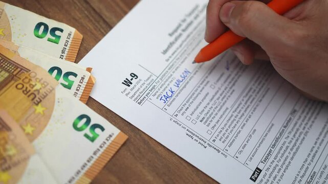 Businessman meticulously writes on W-9 tax declaration related to household office expenses using pen, man works near euro banknotes placed on wooden table slow motion