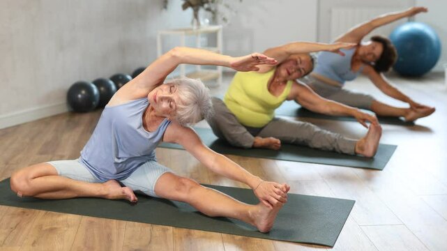 In fitness studio with small group of friends, elderly woman perform Parivritta janu shirshasana. Mature slender women participates in yoga classes.