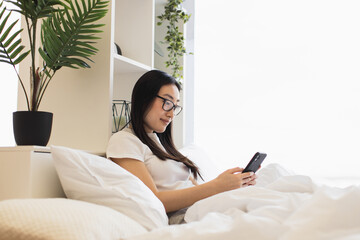 Young Asian woman relaxing on bed using smartphone. Modern bedroom with natural light and greenery creates calming atmosphere.