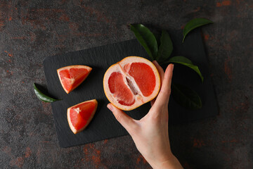 Sliced ripe grapefruit with leaves on board and female hand on dark background, top view