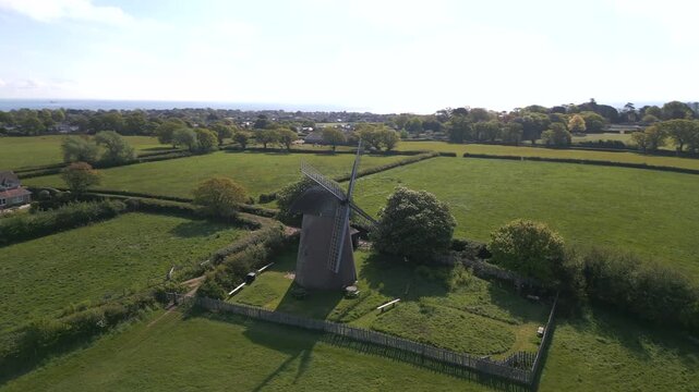 Scenic spinning aerial shot of Bembridge Windmill surrounded by lush green fields, trees, and distant coastal views on a sunny day, Isle of Wight.