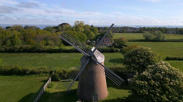 Reverse drone shot flying between the blades of Bembridge Windmill, revealing the historic structure, lush green fields, and countryside views on the Isle of Wight.