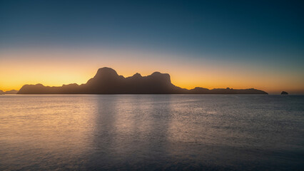 Mysterious tropical sunset. The blue evening sky is gradient illuminated with pink, orange, and yellow. Bizarre silhouettes of mountains on the horizon.Glare on the surface of a calm ocean.Philippines