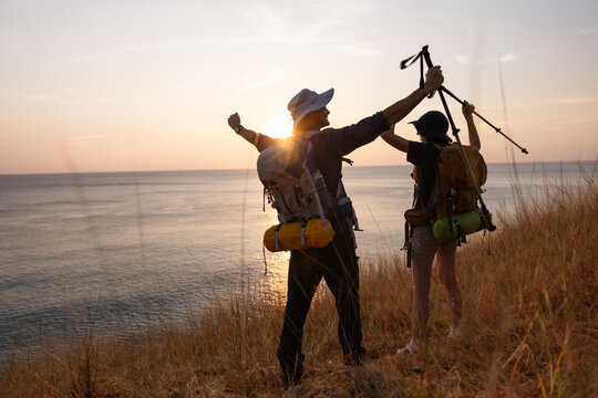 Two hikers celebrating at sunset, standing on a grassy hill overlooking a serene ocean. Equipped with backpacks and trekking poles, they embody adventure, freedom, and the joy of exploration - Powered by Adobe