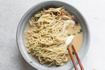 Overhead view of noodles in milk broth in a grey bowl, top view of ramen noodles in creamy chicken broth on a marble countertop 
