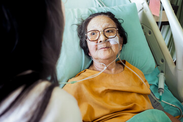 Sick Southeast Asian old female patient in hospital bed with oxygen support wearing eyeglasses seems happy and passionate of being visited by family