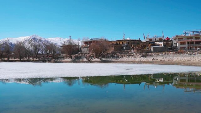Scenic view of partially frozen Nako lake on background of snowy Himalaya mountains in Nako Village, Himachal Pradesh, India. Village houses near lake.