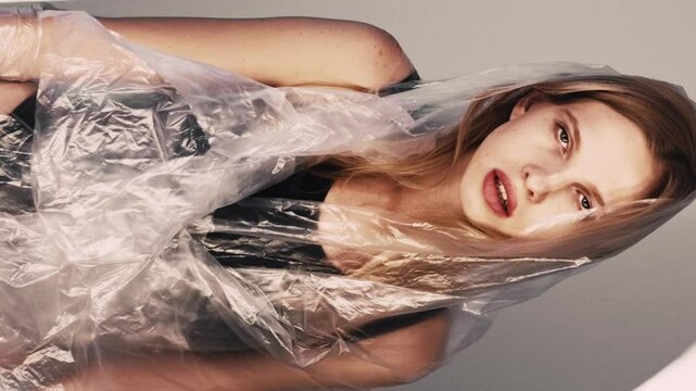 Vertical video. Weight loss. Unhealthy diet. Anorexia problem. Weak underweight woman wrapped in transparent polyethylene film fainting on gray illuminated art.