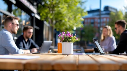 Four young professionals collaborate on laptops at an outdoor cafe table. Flowers and drinks are on the table. Sunny day.