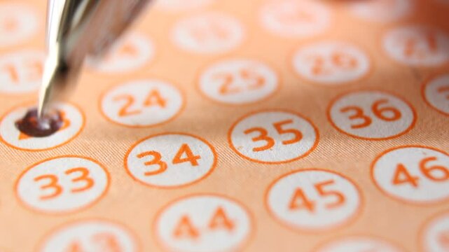 A man marks with a pen numbers 23, 34, 35, 46 on a lottery ticket. The rows of orange numbers in white circles on the orange lottery ticket are arranged diagonally. Close-up horizontal video.