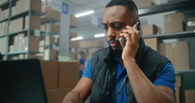 Warehouse retail facility of E-commerce store or post office: African American logistics coordinator talks by phone, uses laptop computer. Sorting center workers carrying cardboard boxes for delivery.