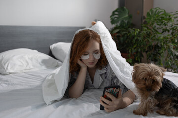 A woman enjoys a cozy morning in bed with her smartphone and a small dog snuggled beside her, enhancing comfort