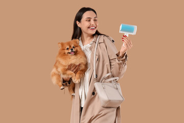 Young woman with lint roller and fluffy dog on brown background