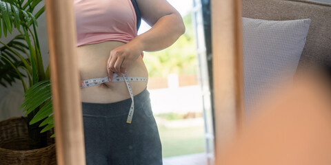 Woman measuring her waist with a tape measure while examining her reflection, focusing on body...
